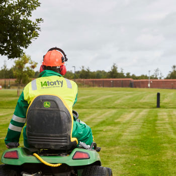 A member of 14forty's outdoor maintenance staff wearing branded fluorescent safety clothing riding a sit-on lawn mower on a large freshly cut lawn.